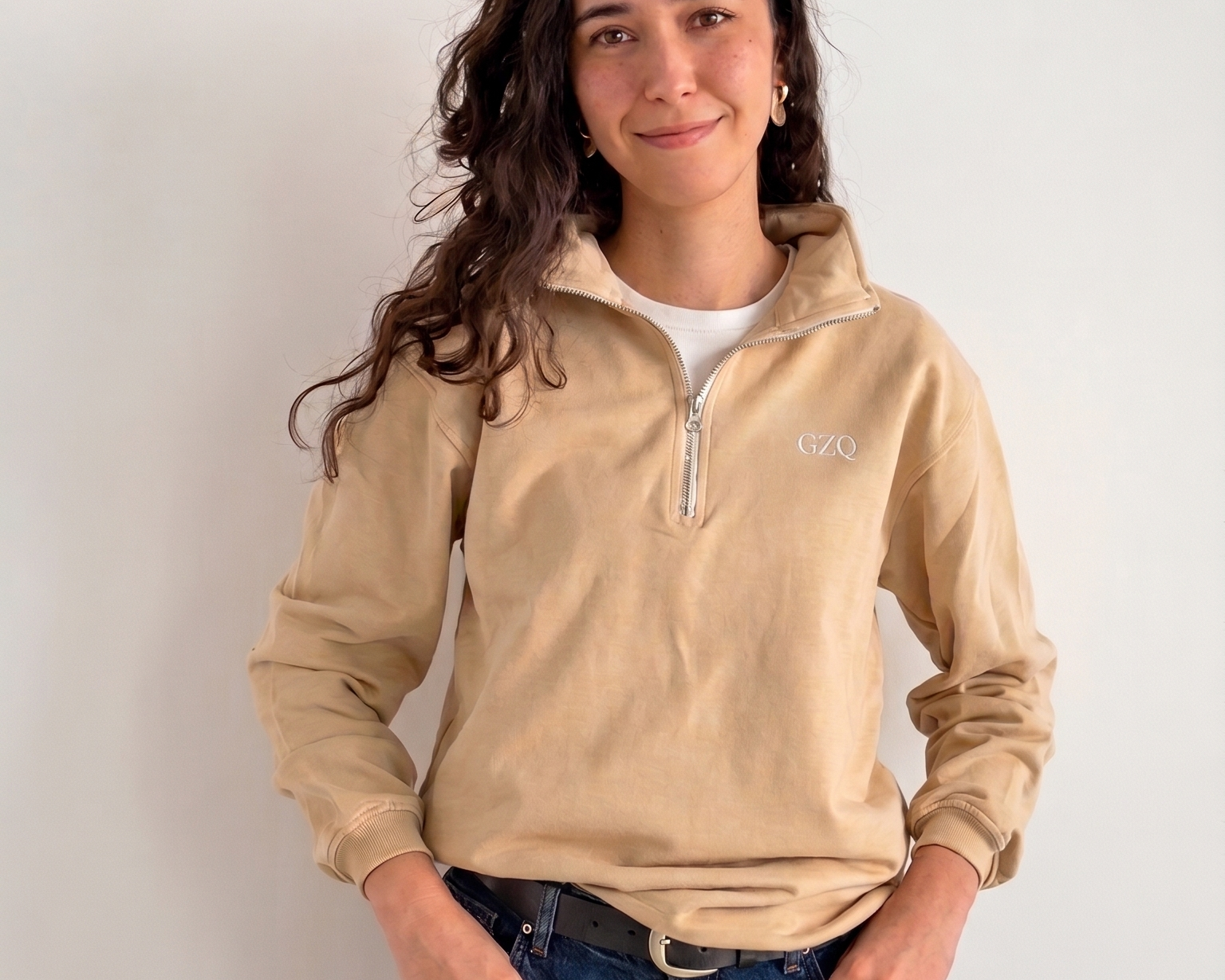 Woman wearing a beige long-sleeve shirt with a logo and blue jeans against a white background