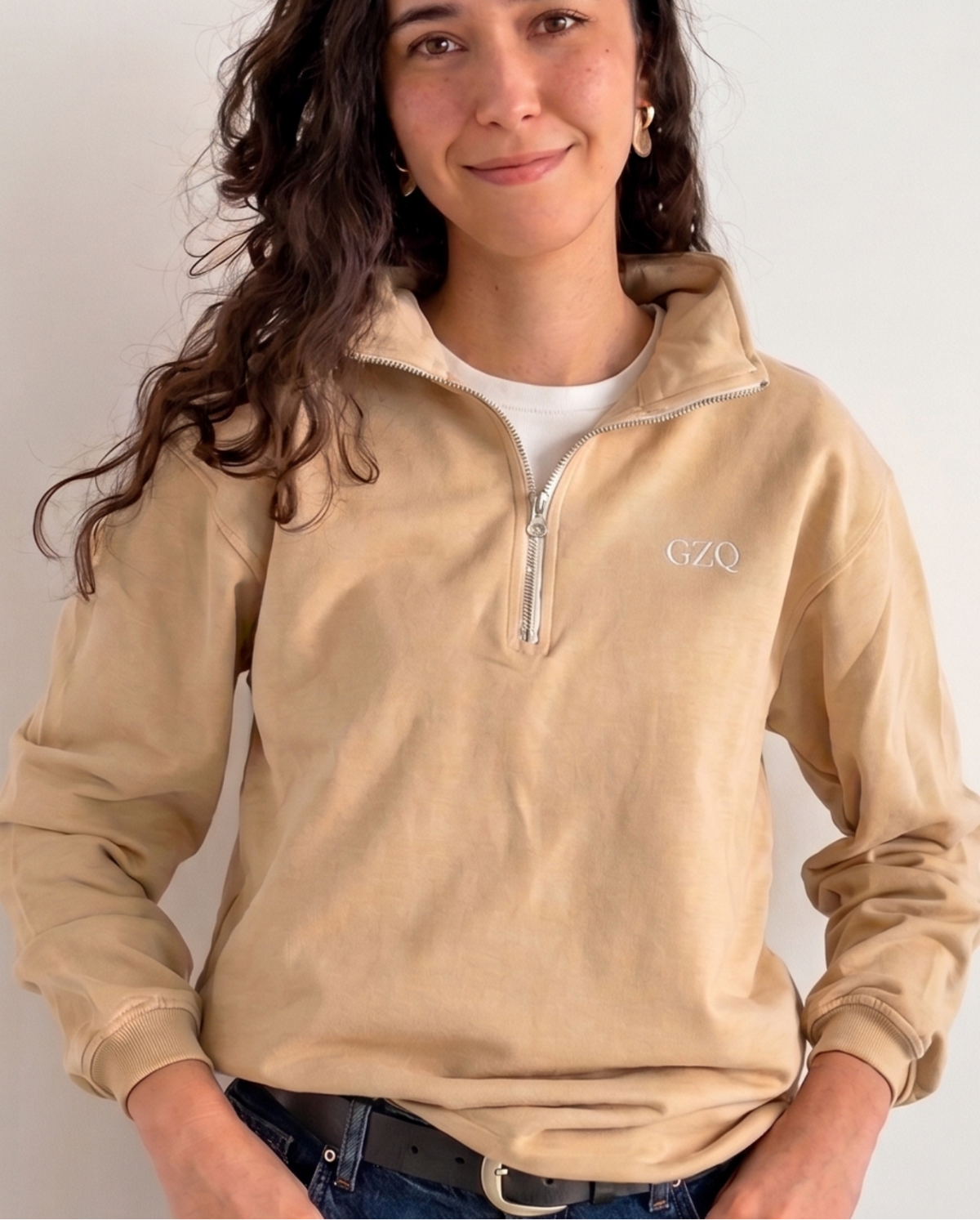 Woman wearing a beige long-sleeve shirt with a logo and blue jeans against a white background